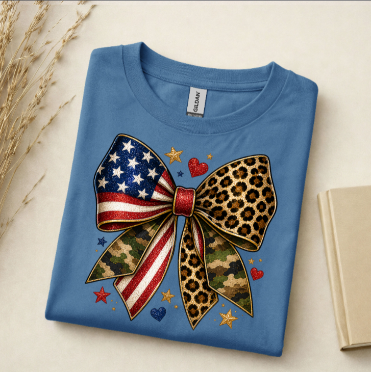 Folded blue t-shirt with a colorful bow design featuring American flag, leopard print, and camouflage patterns